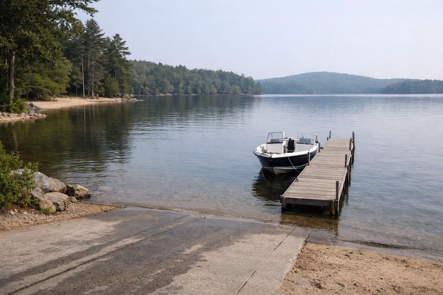 A concrete public boat ramp sloping into a large calm lake, a small motorboat tied at a short dock