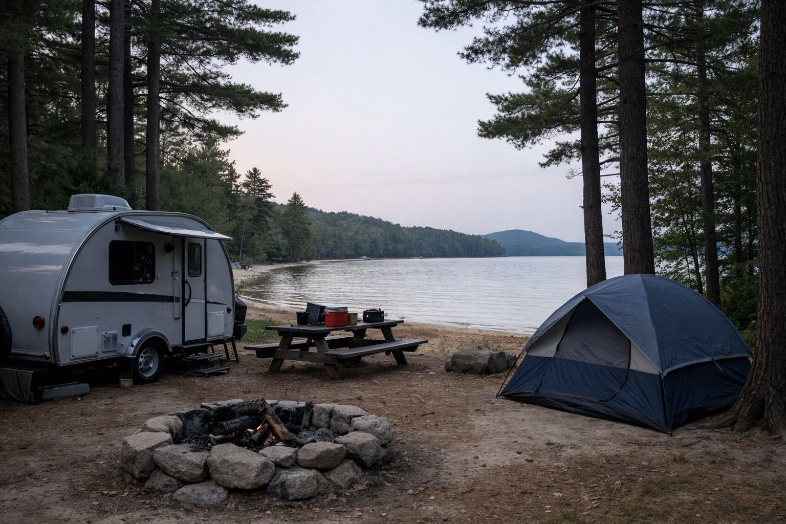 A wooded campsite with a small camper trailer, a tent, a picnic table and a stone fire ring under tall pines