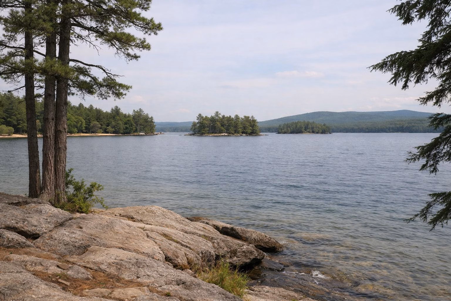 A view across a large lake from a low rocky point with pines, small islands in the distance