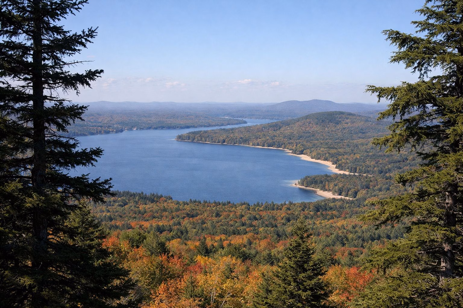 A view from a mountaintop over forested hills in fall color and distant lakes, framed by spruce trees